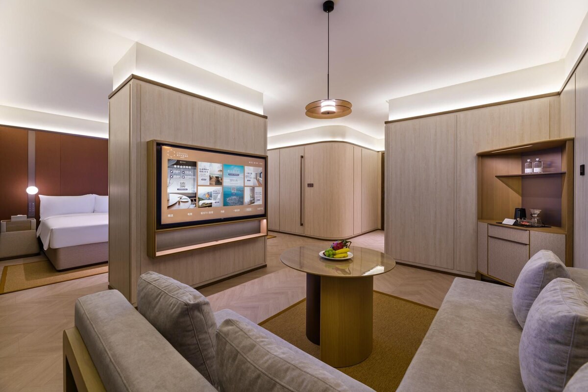 A modern hotel room with a bed, a TV mounted on a wall, a round table with a fruit bowl, and a sofa.