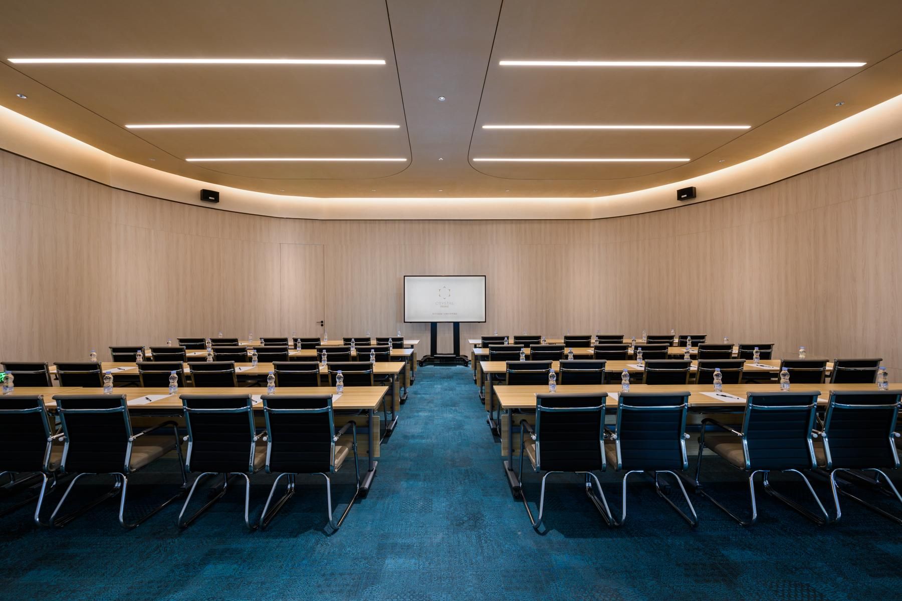 Une salle de conférence avec des rangées de tables et de chaises, un écran de projection à l’avant et un sol recouvert de moquette.