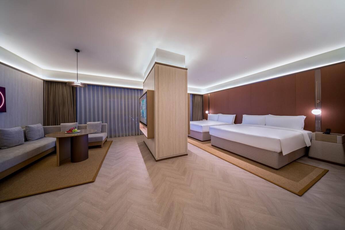 A modern hotel room with a sofa, a dining table, two beds, and a TV.