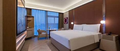 A modern hotel room with a large bed, a desk, and a chair. There is a TV on the left side and a wall-mounted light fixture above the bed.