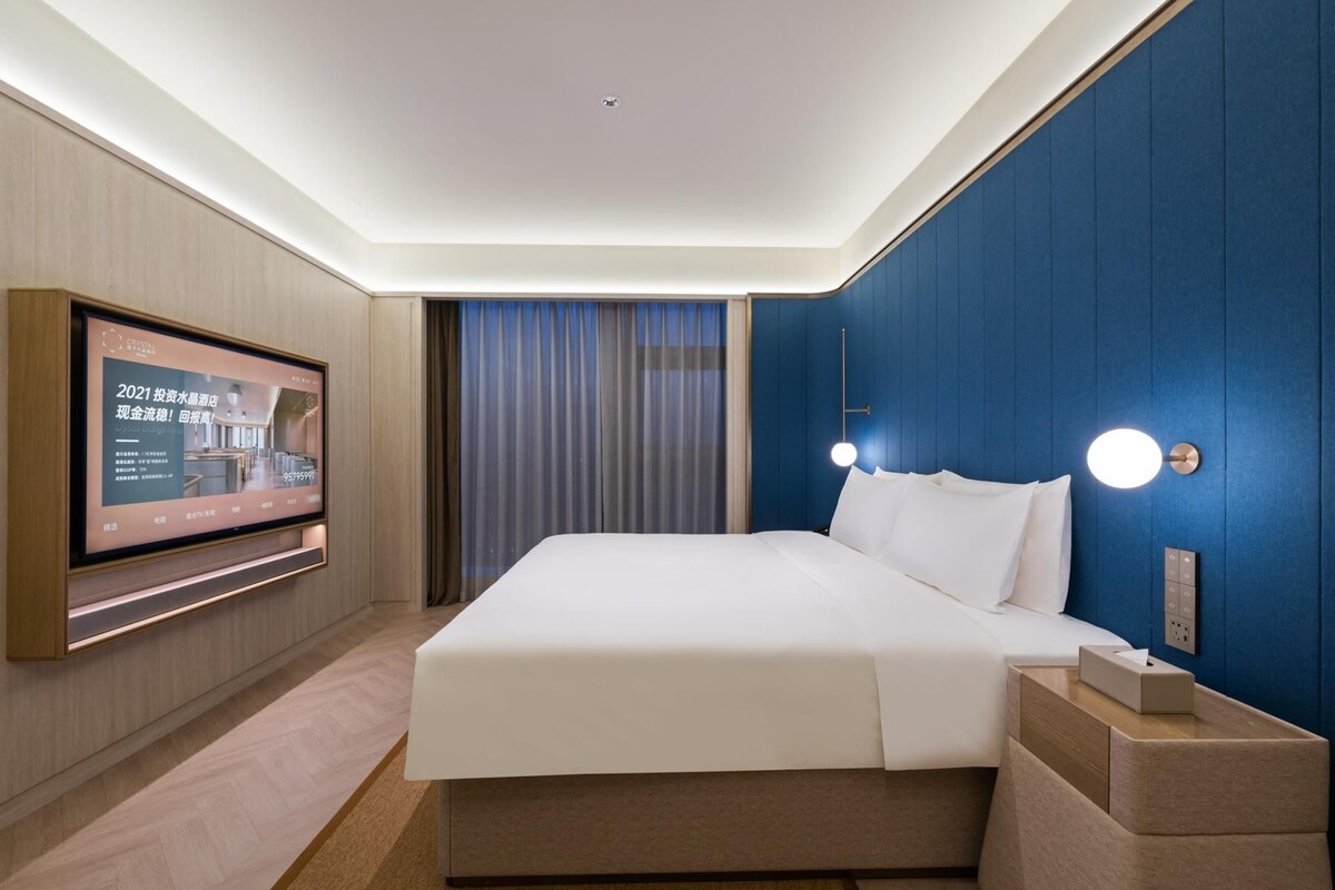 A hotel room with a large bed, a TV mounted on the wall, and a nightstand with a lamp.