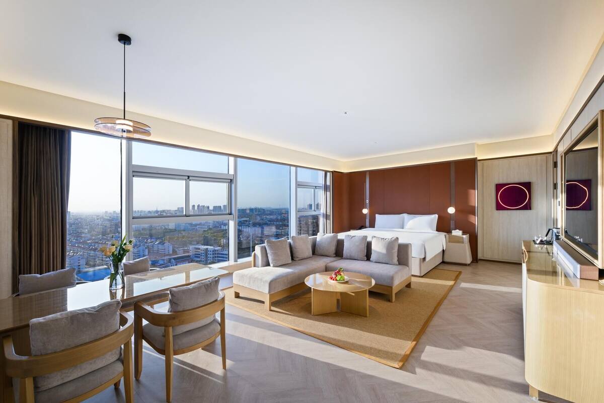 A modern hotel room with a large bed, a sofa, a dining table, and a TV. The room has large windows offering a city view.