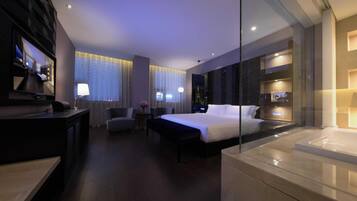A modern hotel room with a large bed, a desk, a chair, a small table with a lamp, and a glass partition.