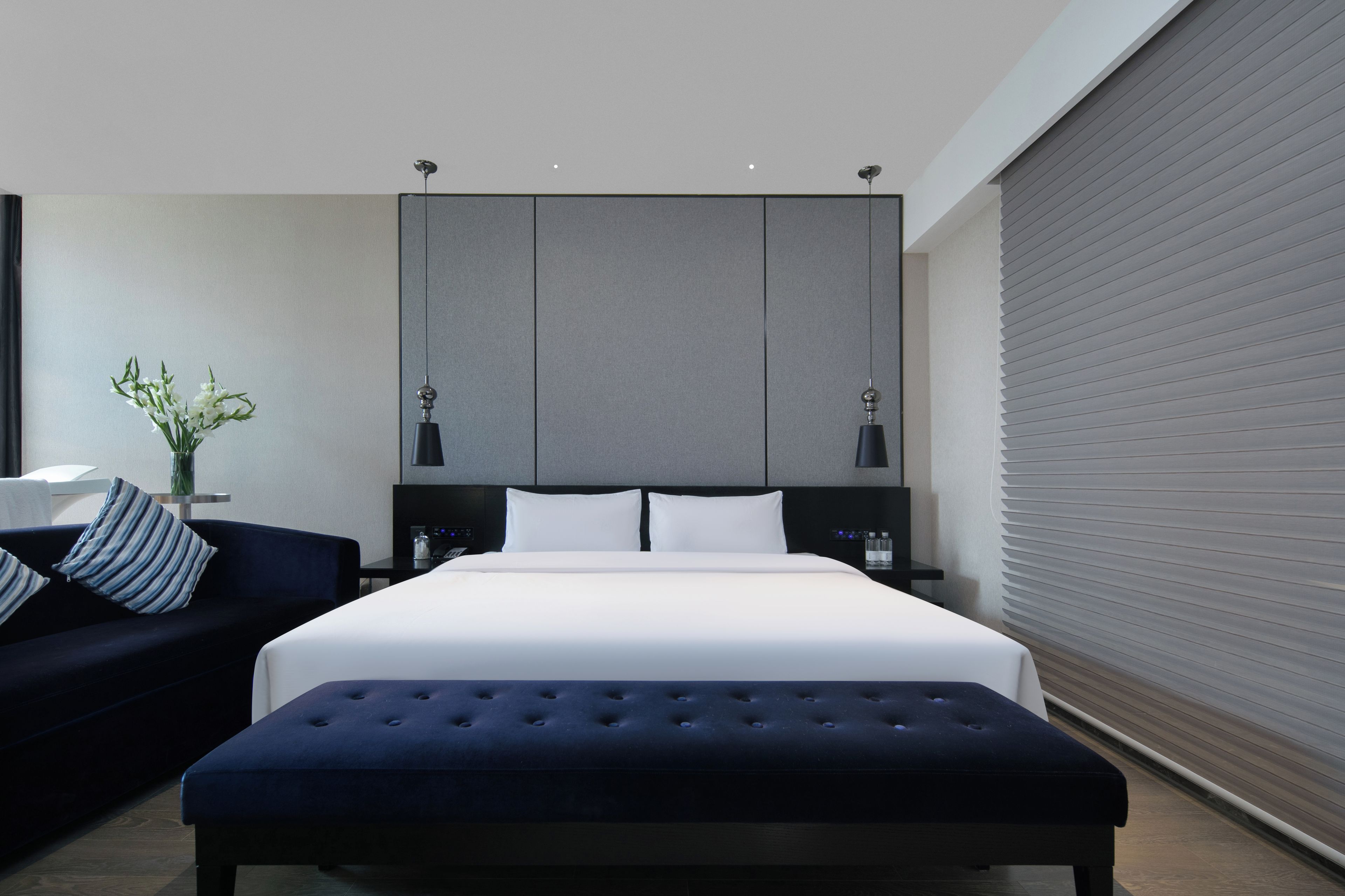 A modern hotel room with a large bed, a dark blue sofa, a small table with a vase, and a large window with blinds.