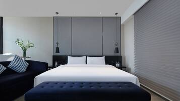 A modern hotel room with a large bed, a dark blue sofa, a small table with a vase, and a large window with blinds.