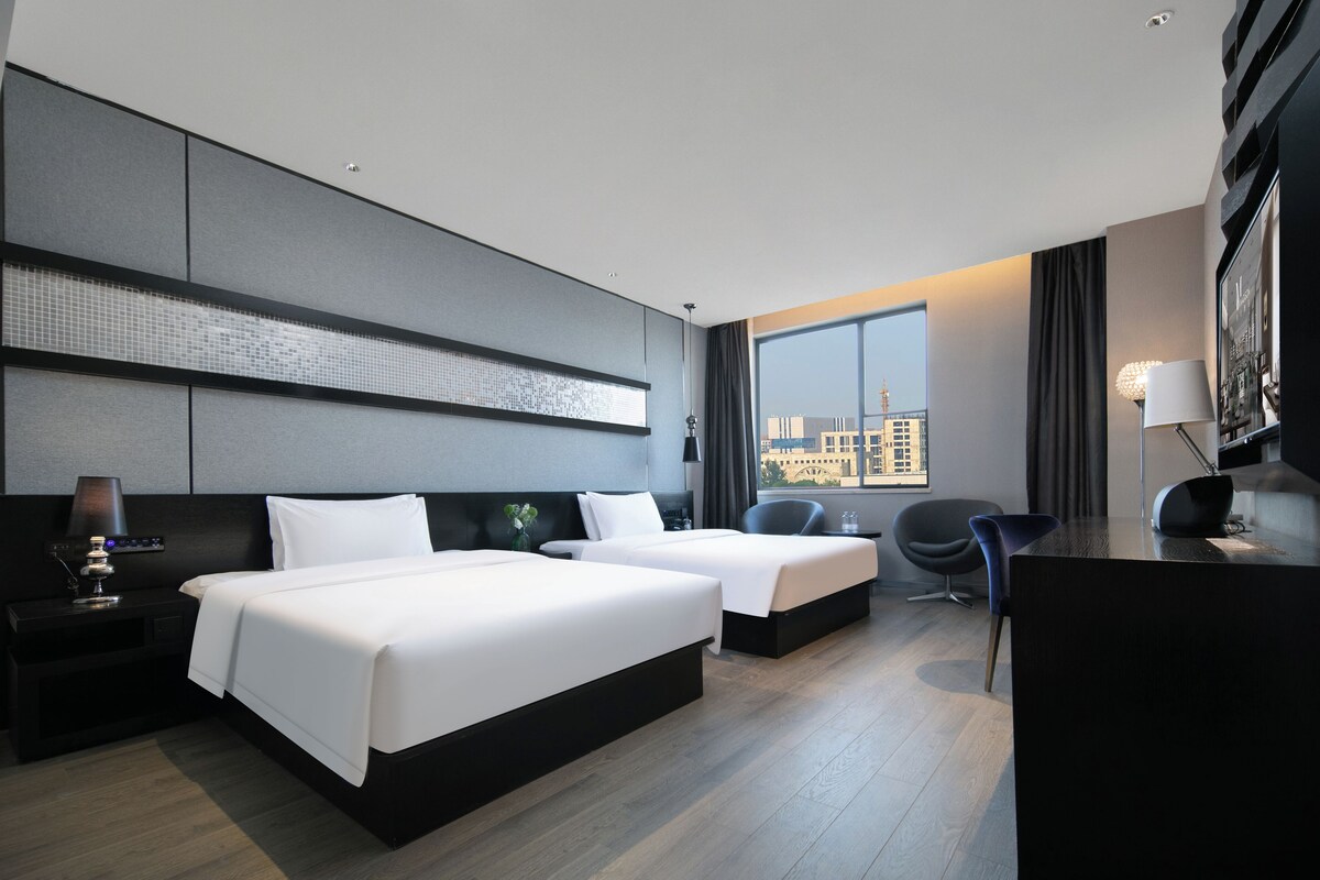 A modern hotel room with two beds, a desk, a chair, and a large window offering a city view.