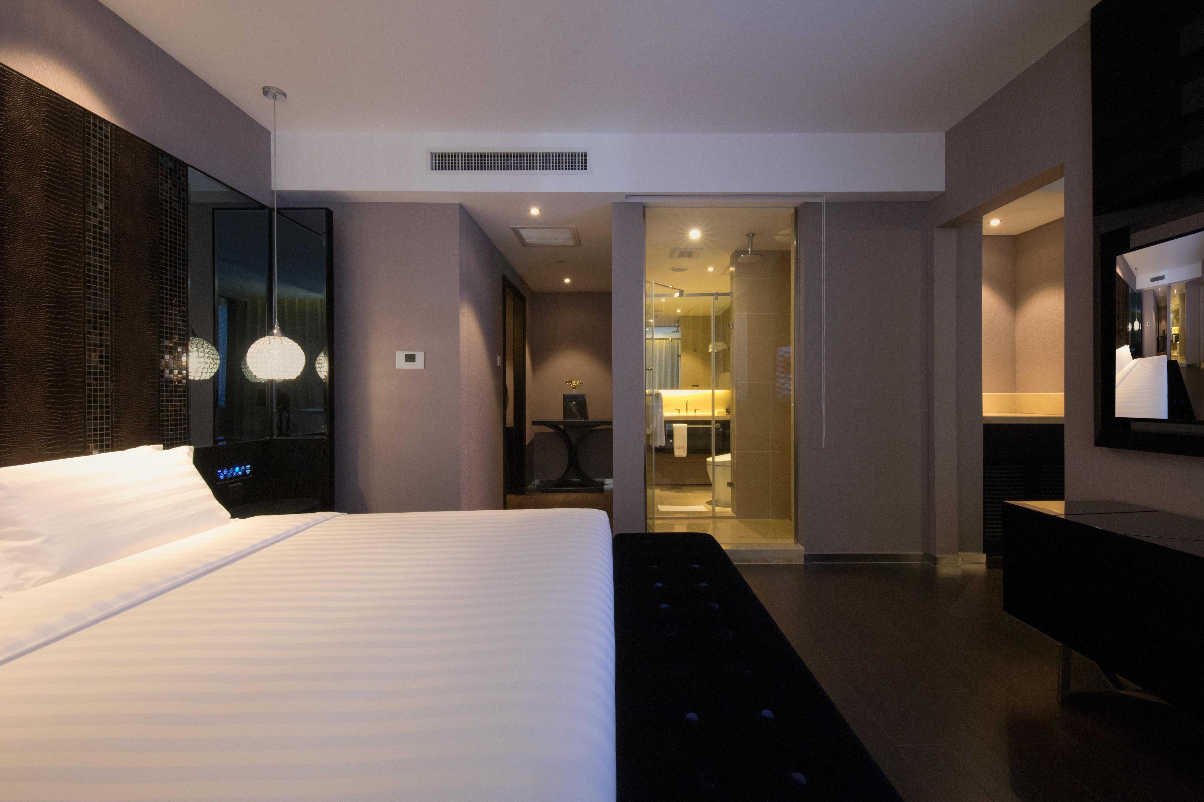 Une chambre d’hôtel moderne avec un grand lit, un banc, un miroir et une salle de bain visible à travers une porte en verre.