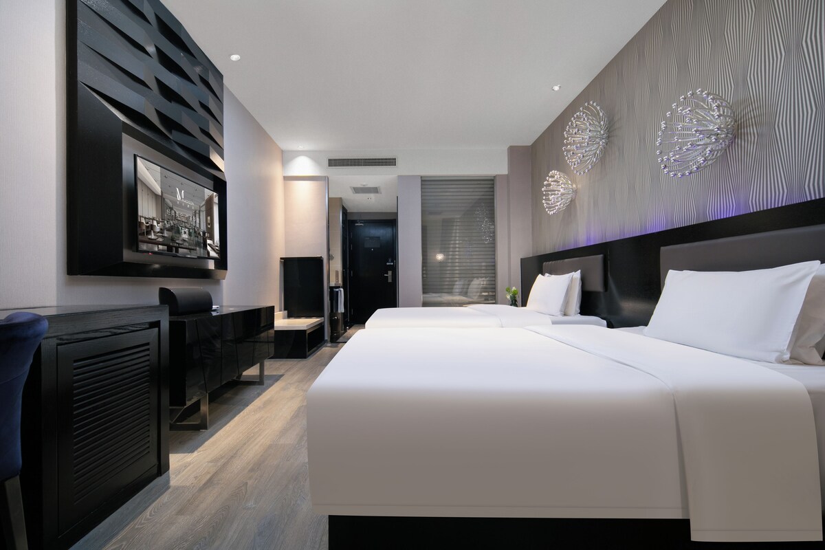 A modern hotel room with a large bed, a desk with a chair, a flat-screen TV mounted on the wall, and decorative wall art.