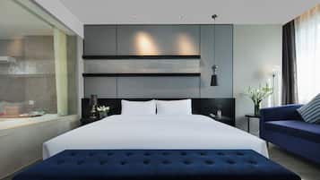 A modern hotel room with a large bed, a sofa, a nightstand, a lamp, and a view of the outside.