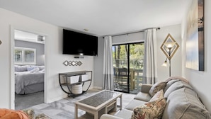 Sala de estar con un sofá, una mesa de centro, un televisor montado en la pared y un ventanal corredizo que da a un espacio exterior.