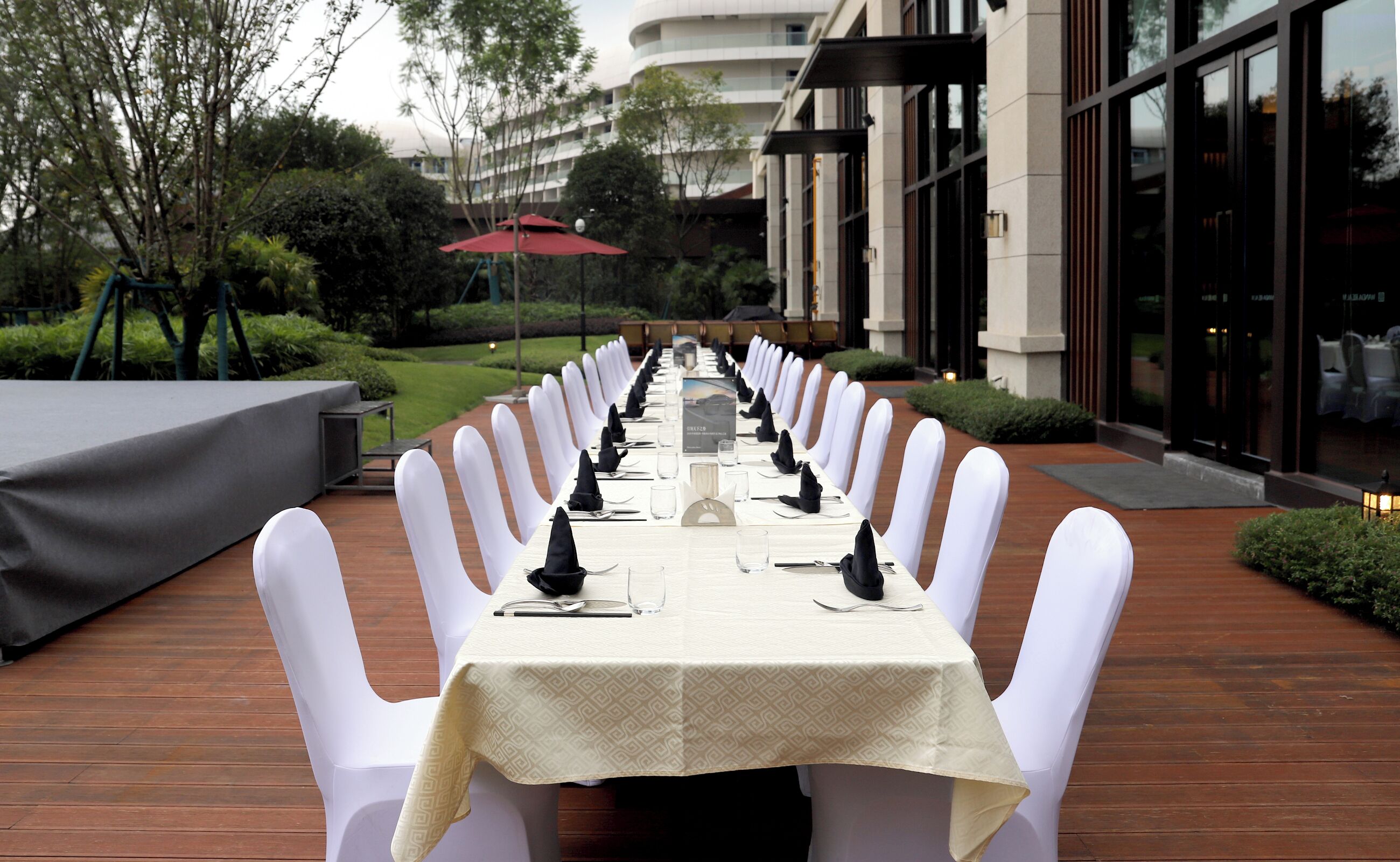 A long dining table set outdoors with white chairs, a covered table, and a red umbrella in the background.