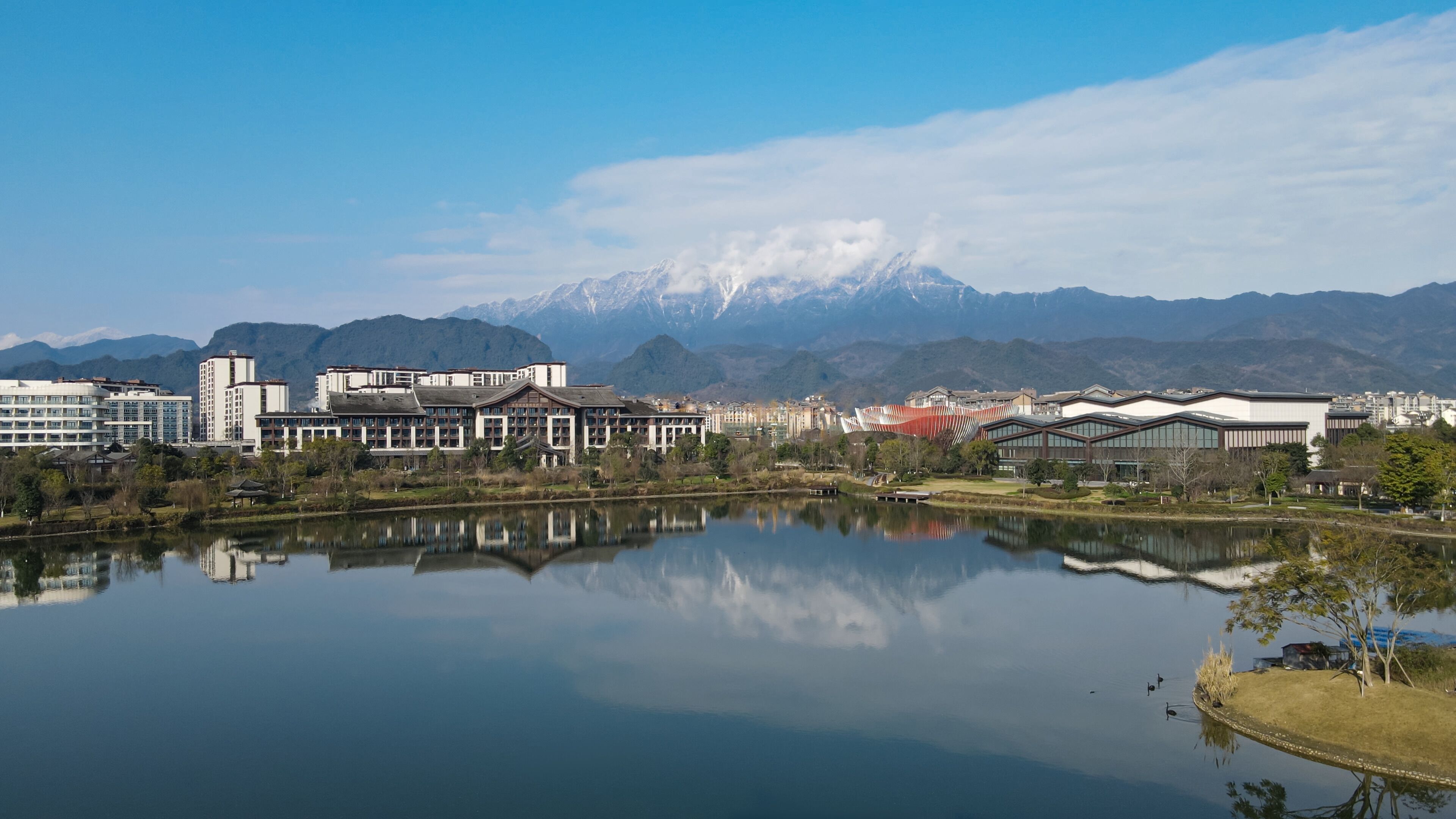 Une vue sur le lac, avec des bâtiments modernes, le reflet de ces constructions dans l’eau, et des montagnes enneigées en arrière-plan.