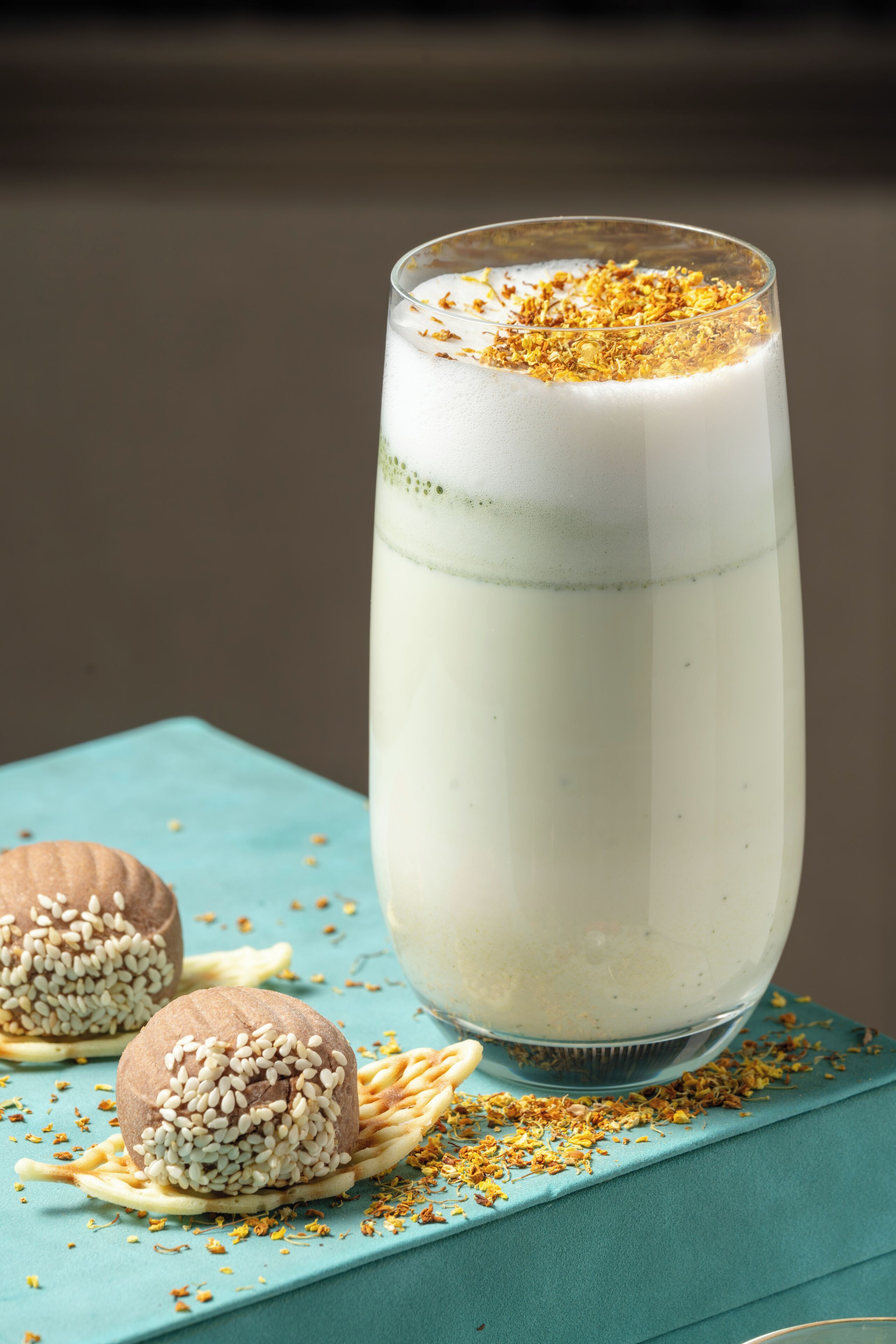 A tall glass of a creamy beverage with a frothy top, placed on a blue surface with two round desserts and scattered crumbs.