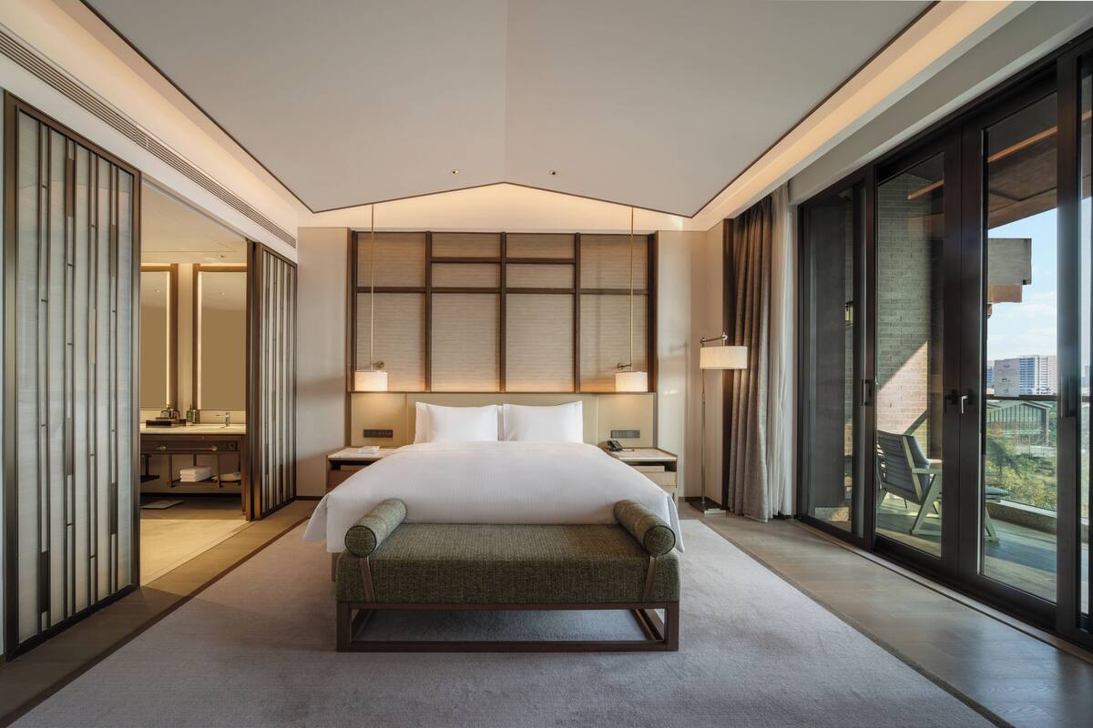 A modern hotel room with a large bed, a sitting area, a bathroom with a mirror, and a view of the city.
