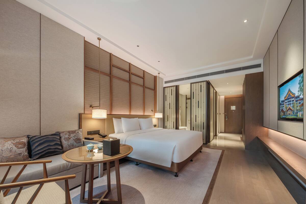 A modern hotel room with a large bed, a sitting area with a table and chairs, and a television on the wall.