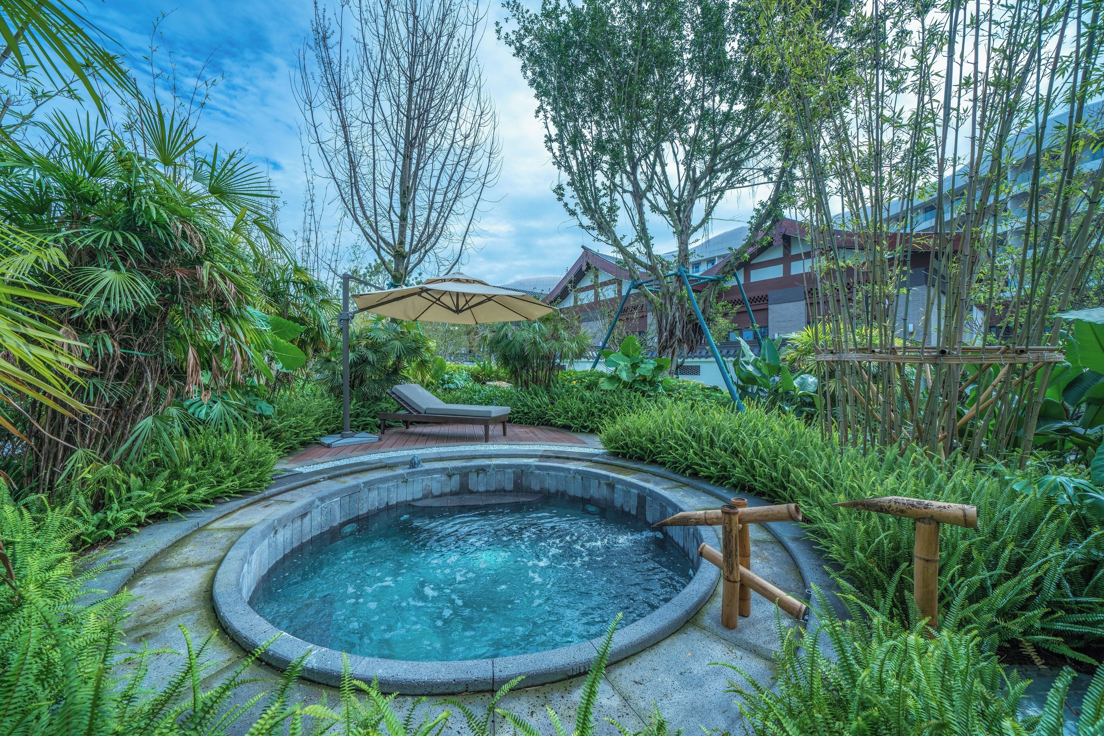 A hot tub surrounded by lush greenery, with a lounge chair and umbrella nearby.