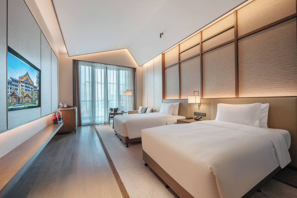 A hotel room with two beds, a desk, a chair, a TV, and a large window with curtains.