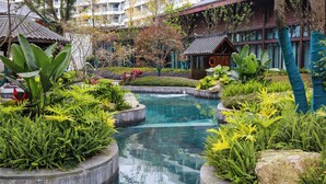 Area piscina con cura del paesaggio, piante tropicali e un edificio tradizionale sullo sfondo.