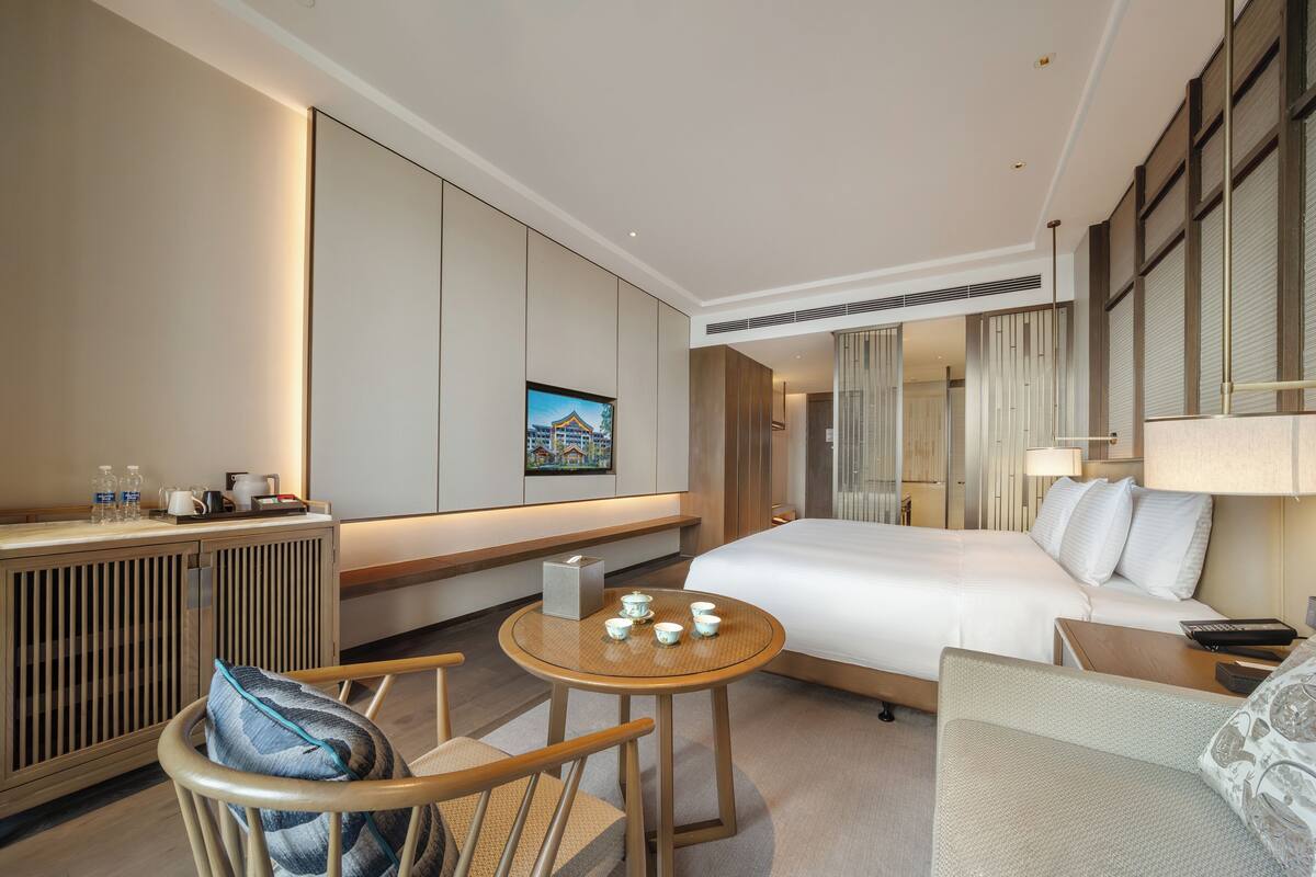 A hotel room with a bed, a seating area with a table, a sofa, a desk with a lamp, and a large window with blinds.
