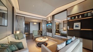 Una moderna camera d'hotel con divano, poltrona e un piccolo tavolino rotondo. C'è una TV montata a parete e una libreria con oggetti decorativi.