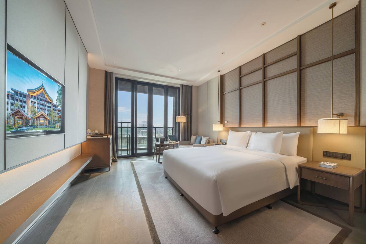 A hotel room with a large bed, a desk, a chair, a TV, and a balcony with a view of a building.