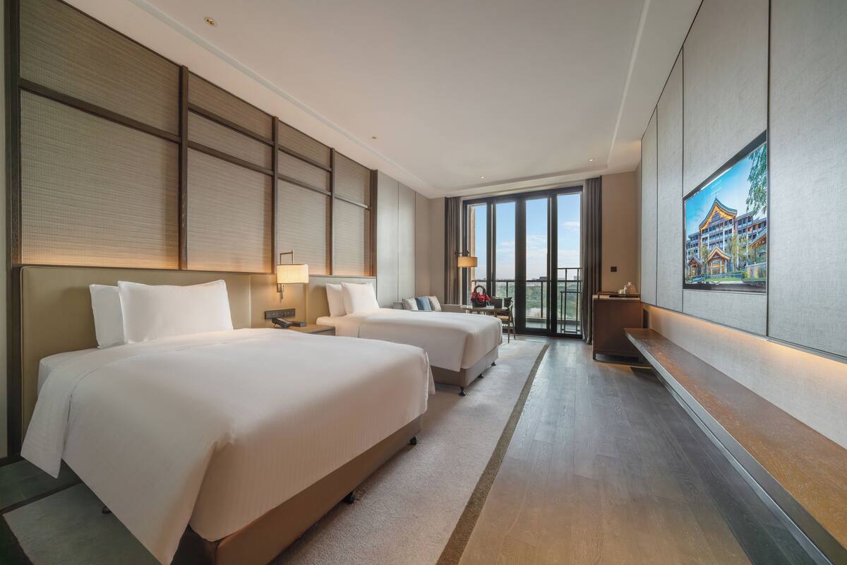 A hotel room with two beds, a desk, a chair, a large window, and a painting on the wall.