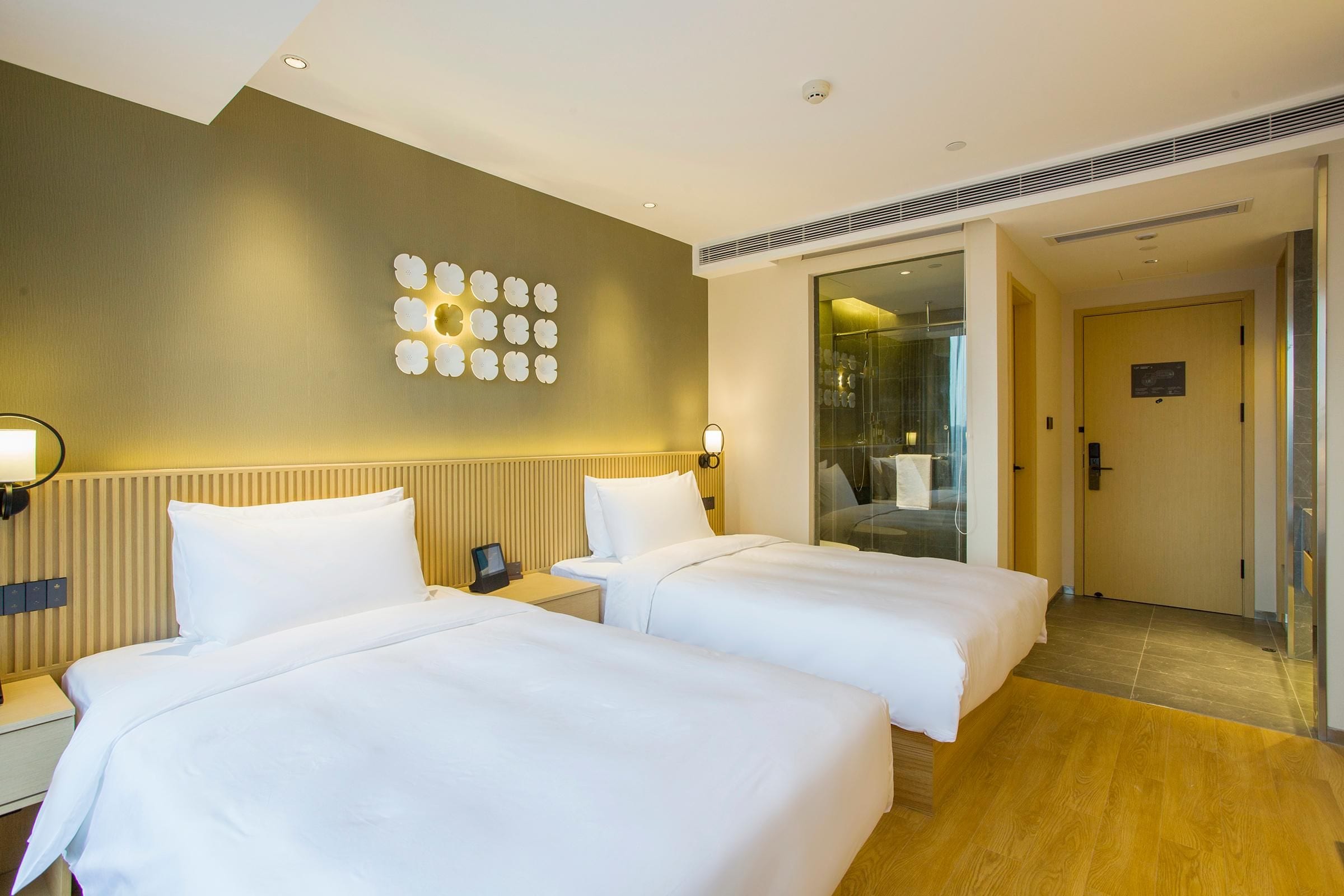 A hotel room with two beds, a wall-mounted light fixture, a glass shower enclosure, and a wooden floor.
