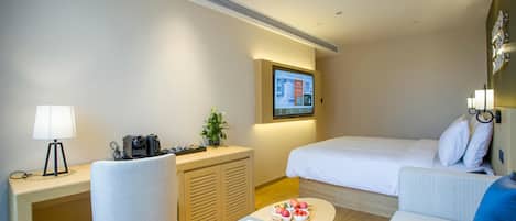 Habitación de hotel con una cama, un escritorio con lámpara, una silla, una mesita con frutas y un televisor fijado a la pared.
