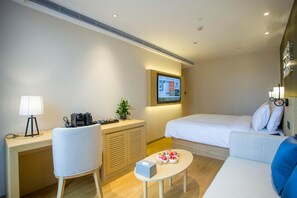 Habitación de hotel con una cama, un escritorio con lámpara, una silla, una mesita con frutas y un televisor fijado a la pared.
