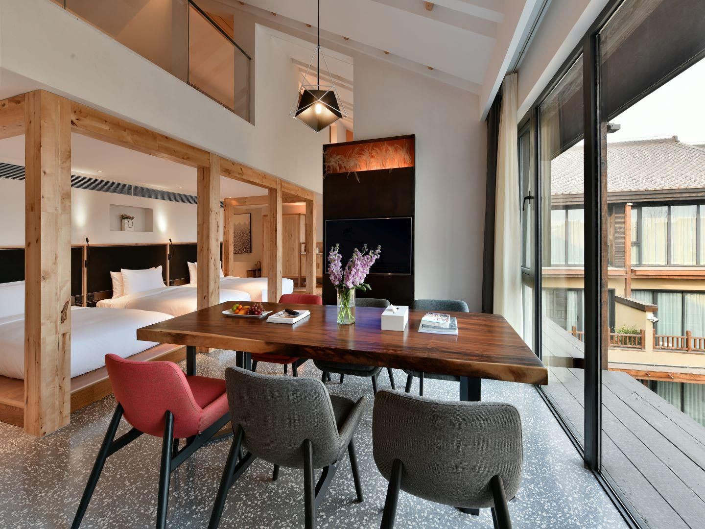 A modern hotel room with a wooden dining table, four chairs, a vase with flowers, and a large window offering a view of the outside.