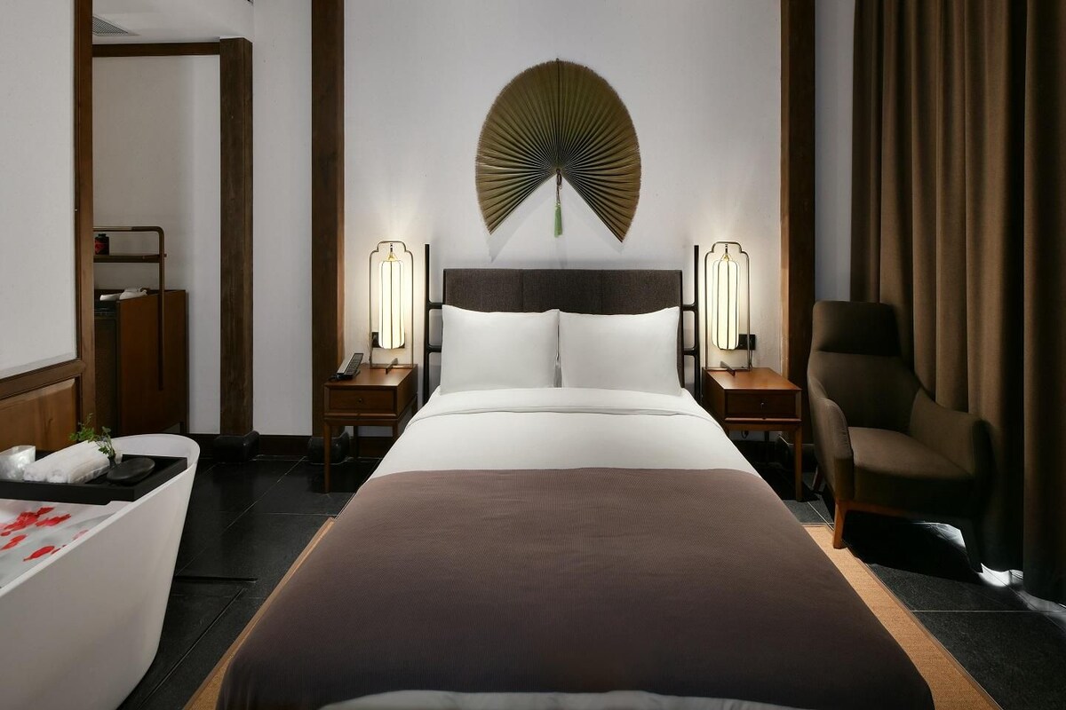 Habitación de hotel con una cama grande, dos mesitas de noche con lámparas, una silla y un adorno en la pared.