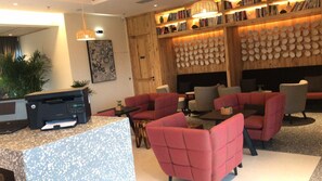 Un hall d'hôtel comprenant un comptoir d'accueil, un espace salon avec des fauteuils à revêtement rouge, une bibliothèque et un mur orné de motifs circulaires.