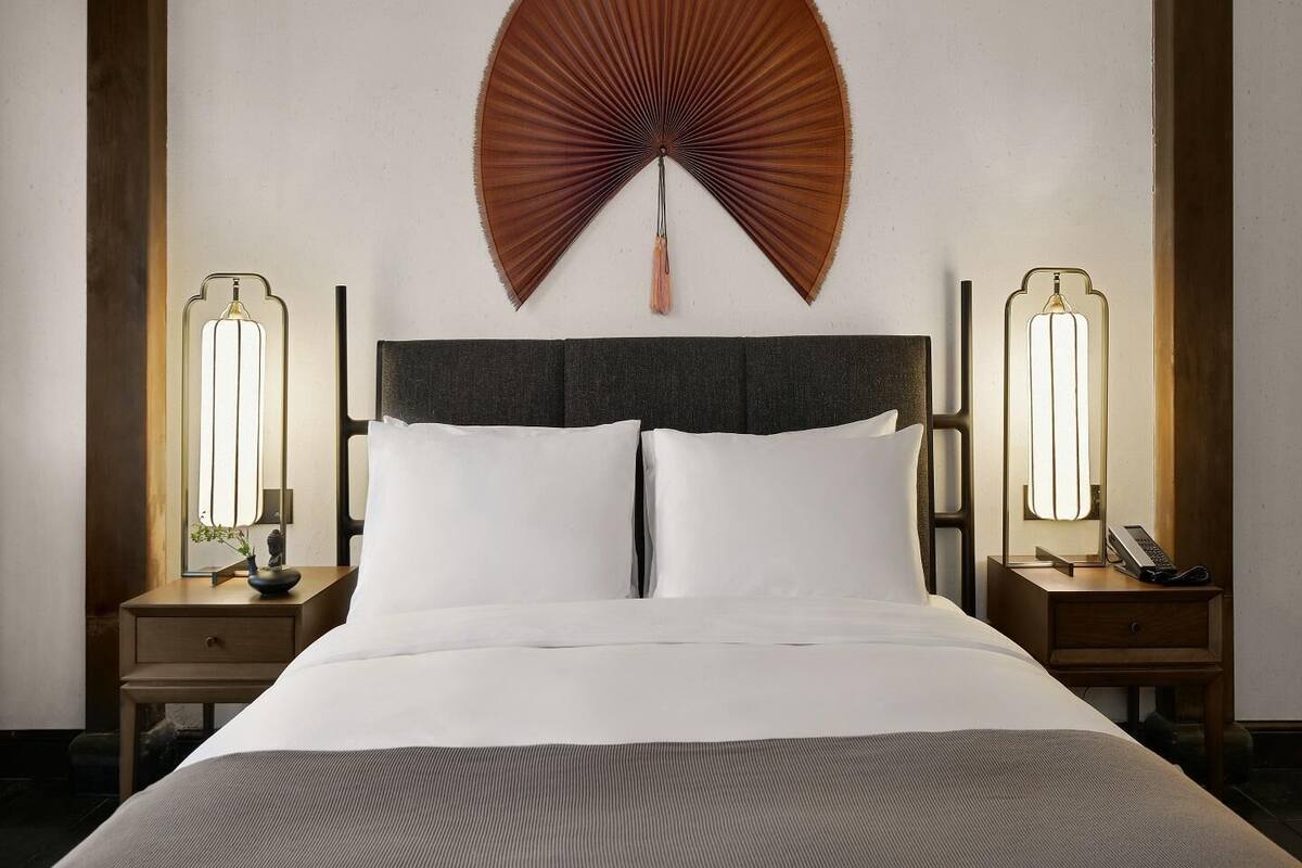 Ein Hotelzimmer mit einem großen Bett, zwei Nachttischen mit Lampen, einem Wandventilator und einem Holz-Kopfteil.