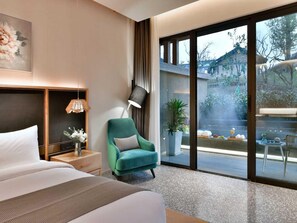 Ein Hotelzimmer mit einem Bett, einem Nachttisch, einer Lampe, einem Sessel und einem großen Fenster mit Blick nach draußen.