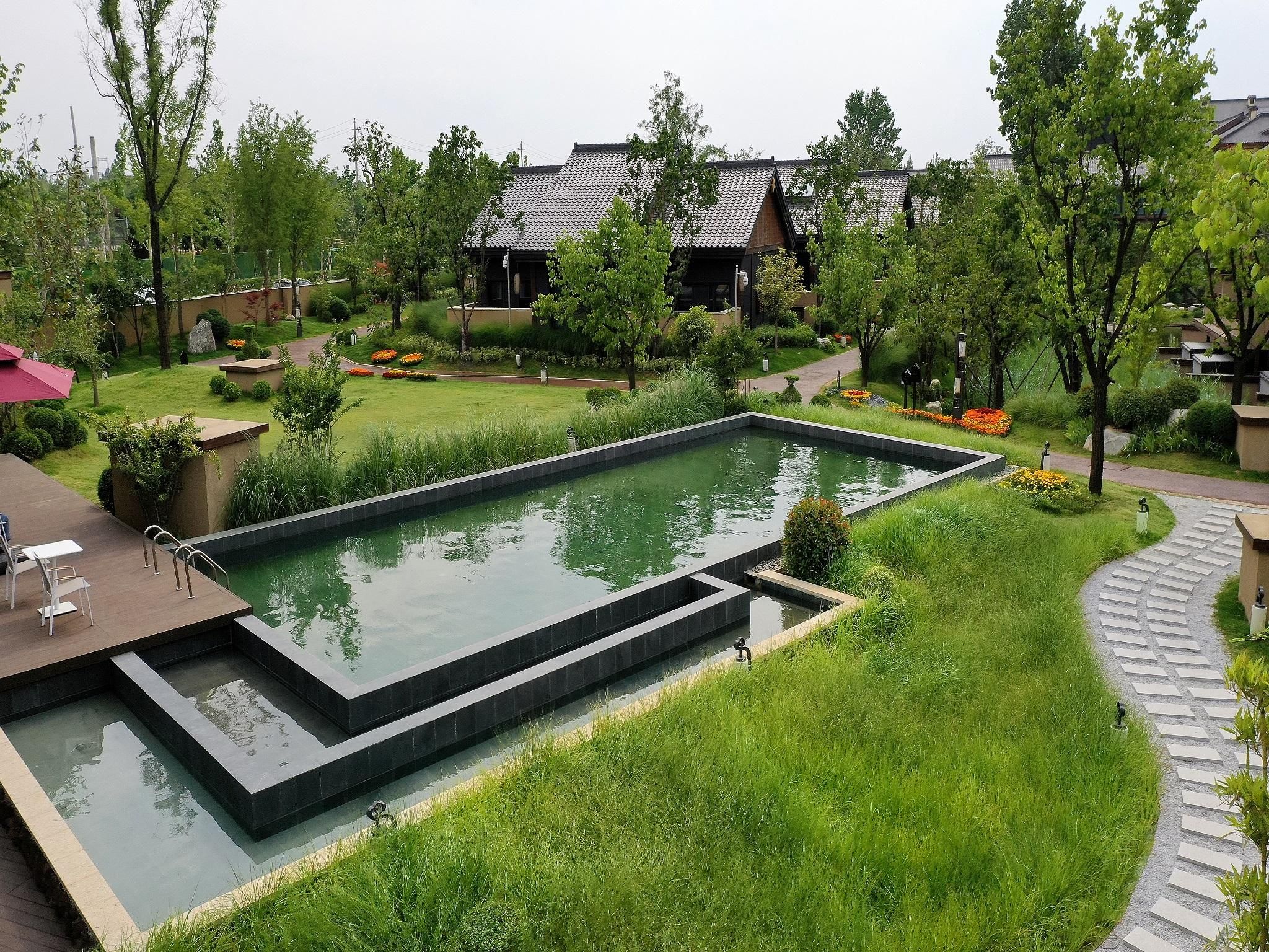 A rectangular pool with a surrounding garden, a pathway, and traditional-style buildings in the background.
