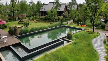 A rectangular pool with a surrounding garden, a pathway, and traditional-style buildings in the background.