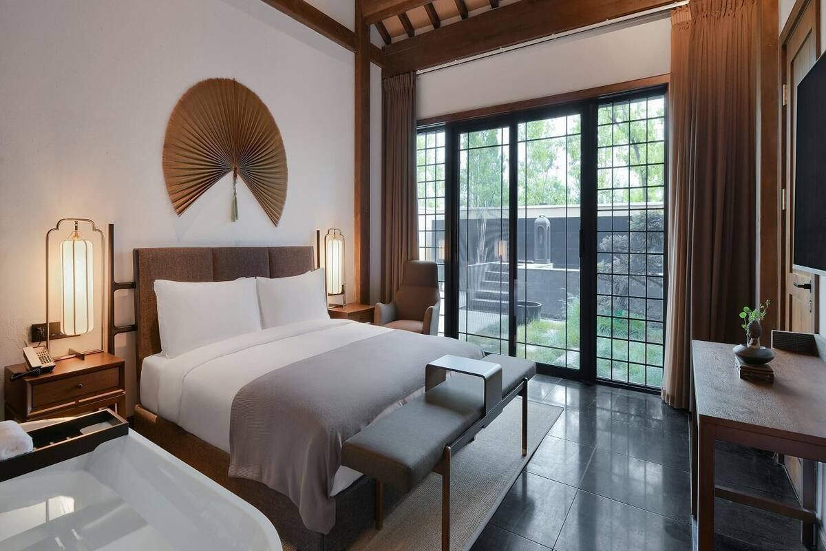 Ein modernes Hotelzimmer mit einem großen Bett, Nachttischlampen, einer Bank, einem Schreibtisch und Blick ins Freie durch große Schiebetüren.