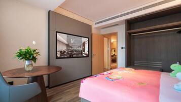 A hotel room with a bed, a desk, a chair, a potted plant, and a view of the hotel's lobby.