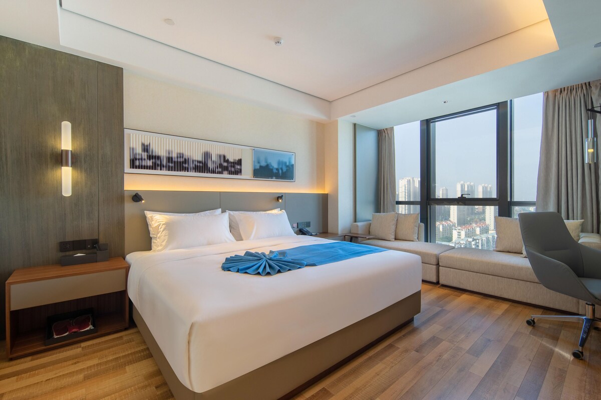 Quarto de hotel moderno com uma cama grande, sofá, escrivaninha e vista da cidade através de janelas amplas.