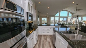A modern kitchen with stainless steel appliances, white cabinetry, and a large window offering a view of the sea. - C'EST FINI - Enjoy superb ocean views & great beach access from this 4,000 plus sqft. stunning home! (Southern Shores)