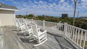 A wooden deck with white railings and rocking chairs, overlooking a coastal area with houses and a clear sky. - C'EST FINI - Enjoy superb ocean views & great beach access from this 4,000 plus sqft. stunning home! (Southern Shores)