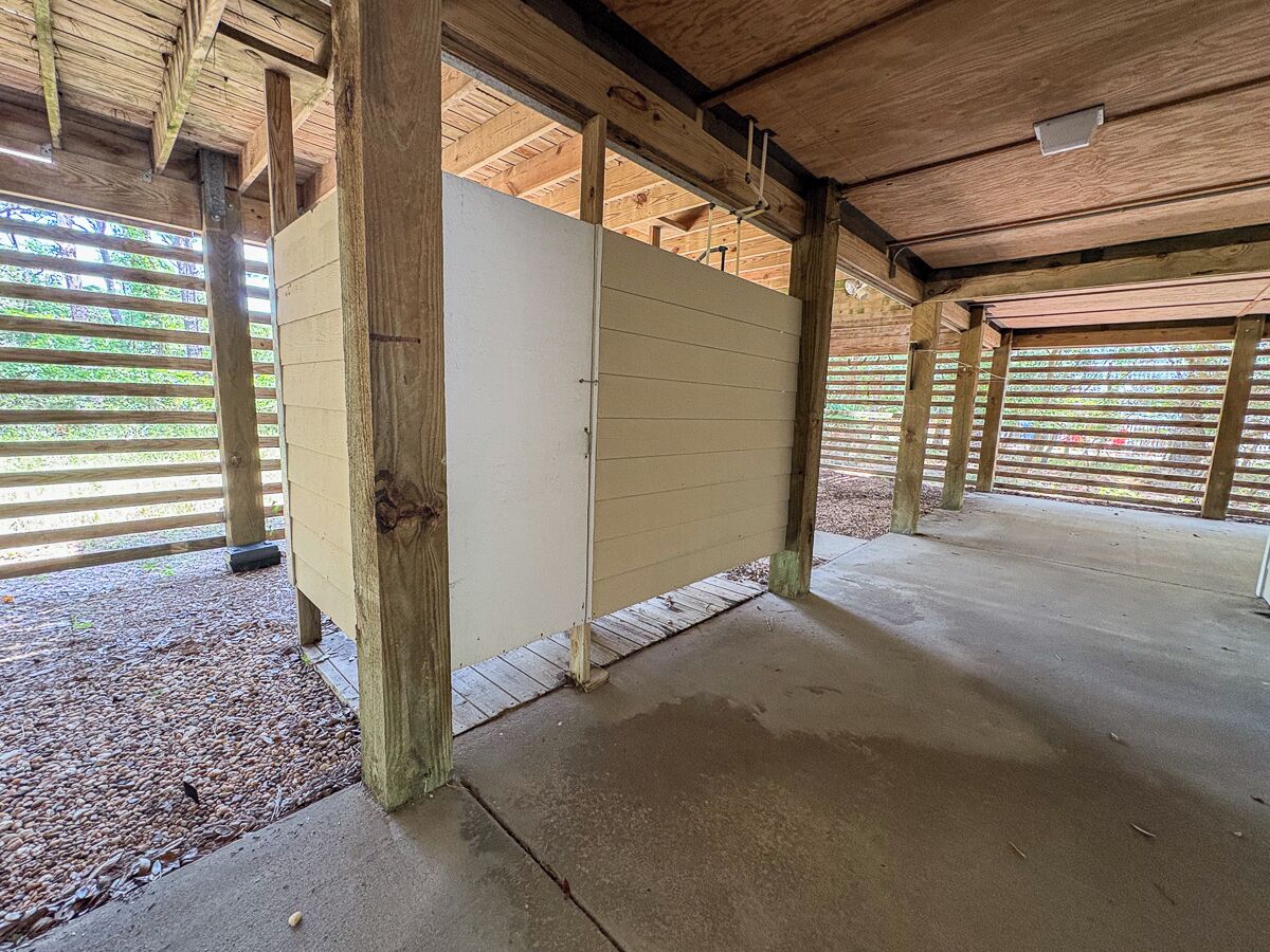 A covered outdoor area with a concrete floor, wooden beams, and a white partition wall.