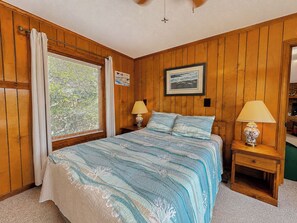 Une chambre avec des lambris en bois, un lit recouvert d’une couvre-lit à motifs floraux bleus et blancs, deux lampes de chevet, une fenêtre avec des stores et un tableau encadré accroché au mur.