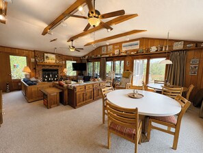 Interior acolhedor de uma cabana com uma mesa de jantar de madeira, área de cozinha com lareira e uma grande janela com vista para o exterior.
