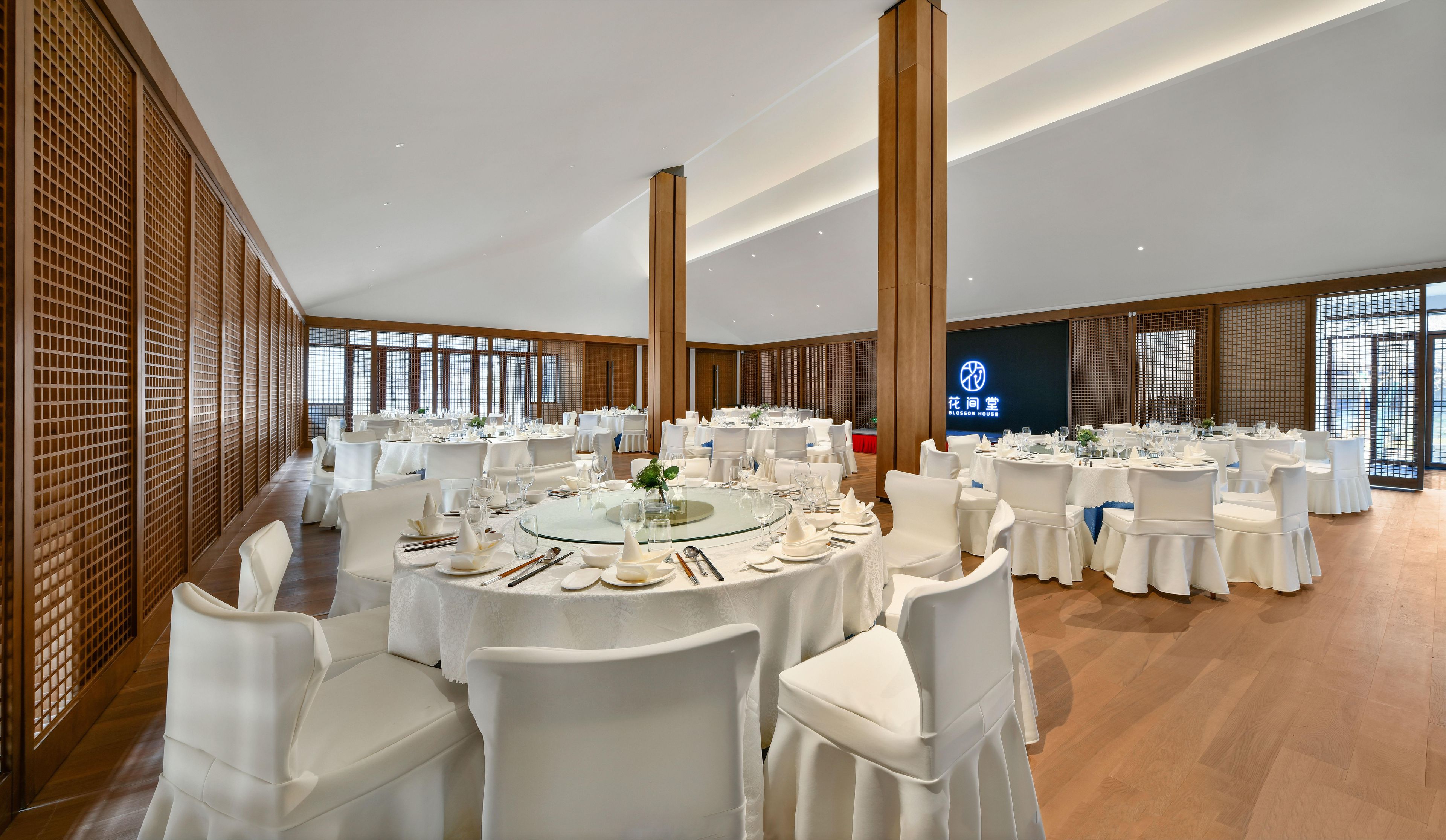A banquet hall with round tables, white chairs, and a large screen displaying the hotel name.