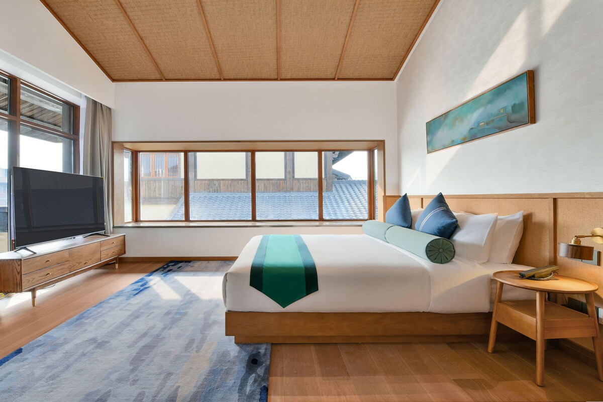 A modern hotel room with a large bed, a TV, a wooden nightstand, and a window with a view of the roof.