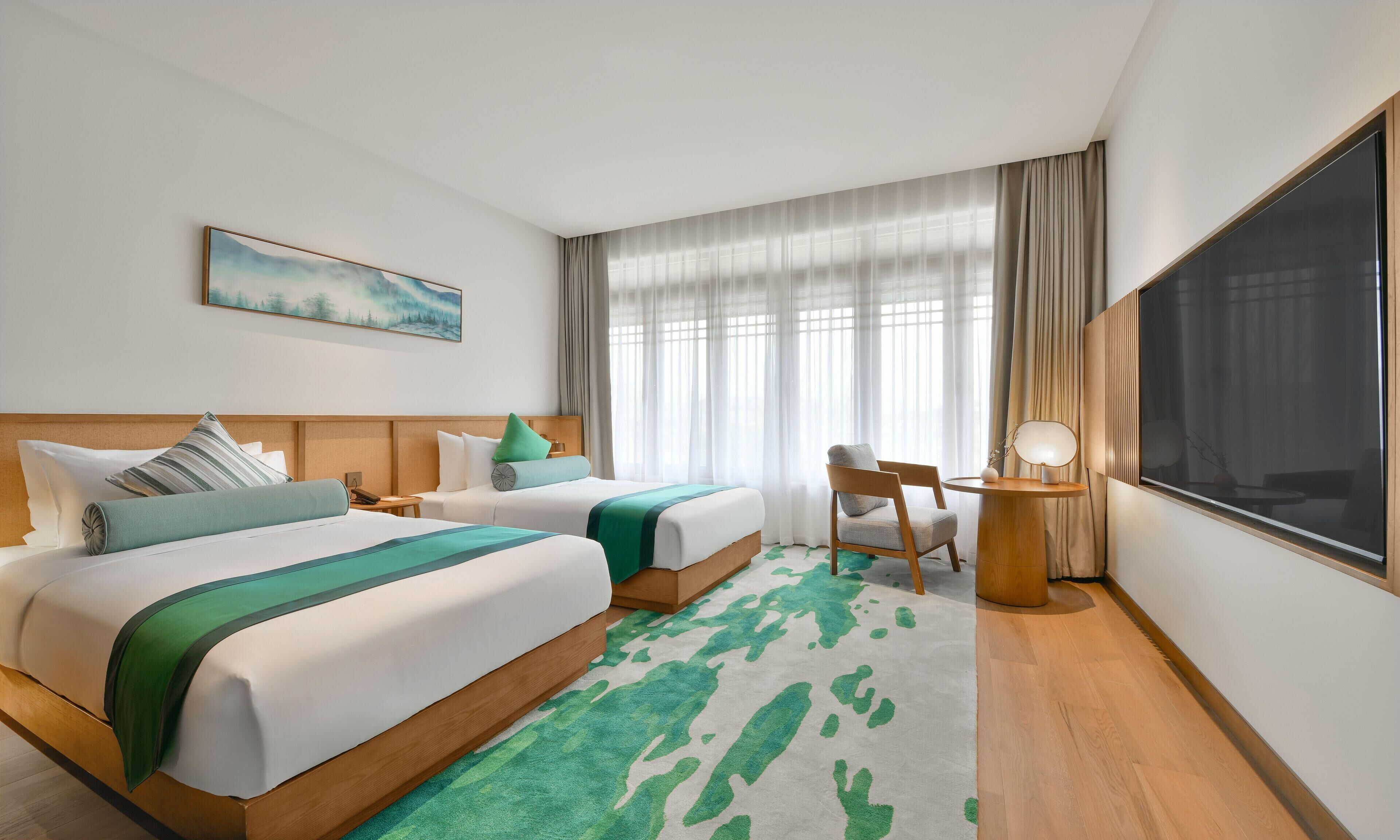 A hotel room with two beds, a desk, a chair, a large window with curtains, and a painting on the wall.