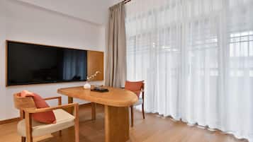 Ett modernt vardagsrum med ett matbord i trä, två stolar, en platt-TV som är monterad på väggen och tunna, genomskinliga gardiner i fönstren.