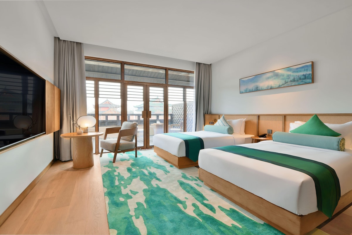 A hotel room with two beds, a desk, a chair, a TV, and a large window with a view of traditional architecture.