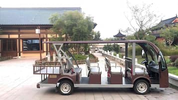 Un rickshaw tradicional con asiento para pasajeros y espacio de carga, estacionado sobre una superficie pavimentada, con un edificio y árboles al fondo.